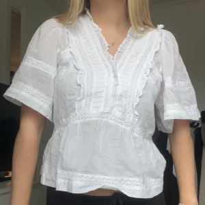 Vit blus med spets från Neo Noir - Superfin vit blus från Neo Noir med volangdetaljer och spets längs v-ringningen och ärmarna. Blusen har korta, vida ärmar och en luftig passform. Perfekt till jeans eller kjol för en fräsch look. Materialet är lätt och känns som bomull. Prislappen kvar. Nypris 600kr