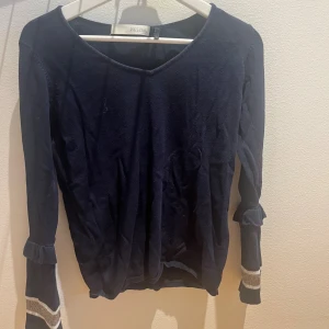 Mörkblå stickad tröja från Pieszak - Mörkblå stickad tröja från Pieszak med långa ärmar och volangdetaljer vid ärmsluten. Ärmarna har även en glittrig rand i silver och vitt. Tröjan har en rundad halsringning och är mjuk och skön, perfekt för kyligare dagar.