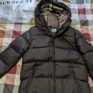 Svart pufferjacka från Burberry - Svart pufferjacka från Burberry med ikoniskt rutigt foder i huvan och insidan. Jackan har huva, dragkedja och knappar med Burberry-logga, samt patch med Burberry London England på ärmen. Perfekt för kalla dagar med sin fluffiga och quiltade design.