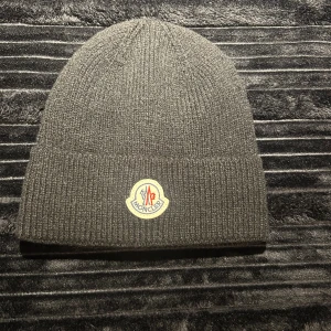 Svart Moncler mössa i ull - Säljer en stilren grå mössa från Moncler med klassisk ribbstickad design och broderad logotyp framtill. Mössan är tillverkad i mjuk ull och passar perfekt för kyliga dagar. Enkel att matcha med olika outfits och ger en exklusiv touch till din look.