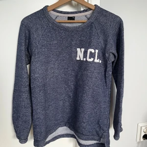 Blå sweatshirt från Gina Tricot XS - Mysig blå sweatshirt från Gina Tricot i storlek XS. Tröjan har trycket 'N.C.L.' på bröstet, rund halsringning och långa ärmar. Perfekt för en chill och avslappnad stil. Materialet är mjukt och skönt, troligtvis bomullsmix.