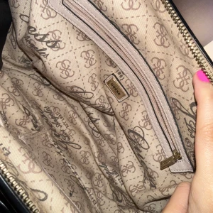 Beige rosa  handväska från Guess - Snygg beige handväska från Guess  mönster, svarta handtag och kedjedetalj framtill. Väskan har en dekorativ rosett och dragkedja upptill. Insidan är mönstrad med Guess-loggor och har en innerficka med dragkedja. Aldrig använd stått bara i skåpet. 