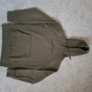 Olivgrön hoodie från Peak Performance - Nypris 1300 kr.  Snygg olivgrön hoodie från Peak Performance med svart logga på bröstet. Tröjan har huva med snörning, magficka och ribbade muddar vid ärmslut och nederkant. Perfekt för chill eller streetwear.