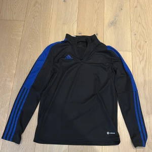 Svart och blå Adidas half zip tröja - Svart långärmad Adidas-tröja med blå detaljer och klassiska ränder längs ärmarna. Tröjan har en half zip framtill och är tillverkad i ett sportigt, syntetmaterial. Perfekt för träning eller chill dagar. Adidas-logga på bröstet och Aeroready-tryck nedtill.