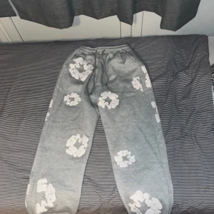 Grå mjukisbyxor Denim Tears med blommor - Grå mjukisbyxor från Denim Tears med vita blommönster över hela byxorna. Byxorna har resår och snörning i midjan för skön passform. Perfekta för chill dagar och streetwear-stil.