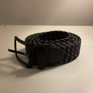 Svart flätat bälte med metallspänne - Snyggt svart flätat bälte med robust metallspänne. Bältet har en klassisk design med vävd struktur och detaljer i svart konstläder vid spännet. Passar perfekt till jeans eller chinos för en stilren look.