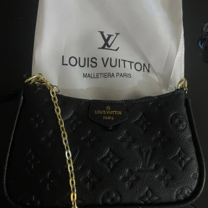 Svart Louis Vuitton axelväska i skinn - Snygg svart axelväska från Louis Vuitton i präglat skinn med klassiskt monogrammönster. Väskan har guldfärgade metalldetaljer, kedjerem och medföljande justerbar axelrem. Perfekt storlek för det viktigaste och riktigt lyxig känsla.