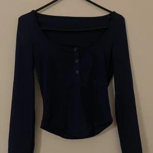 Mörkblå ribbad långärmad topp - Säljer en mörkblå ribbad topp med lång ärm och rundad halsringning. Toppen har fyra knappar framtill som snygg detalj och är figurnära i modellen. Perfekt att styla med jeans eller kjol för en enkel men trendig look.