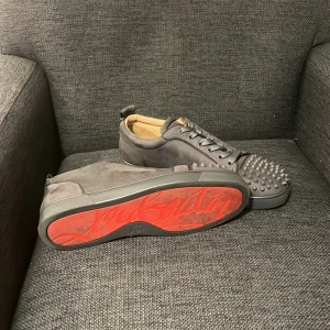 Christian Louboutin grå sneakers - Säljer ett par grå sneakers från Christian Louboutin med ikonisk röd sula och coola nitar på tån. Skorna har snörning och är tillverkade i mocka med rund tå. Insidan är beige och de har en platt sula. Perfekt för dig som vill sticka ut med exklusiv stil.