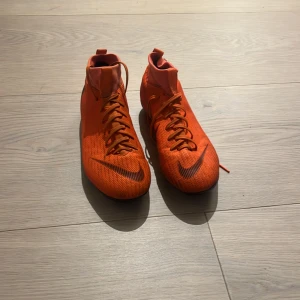 Nike Mercurial orange fotbollsskor - Säljer ett par Nike Mercurial fotbollsskor i en riktigt snygg orange färg med svarta detaljer och logga. Skorna har snörning, högre skaft och platt sula med dobbar för gräsplan. Tillverkade i syntetmaterial för lätt känsla och bra grepp på planen.
