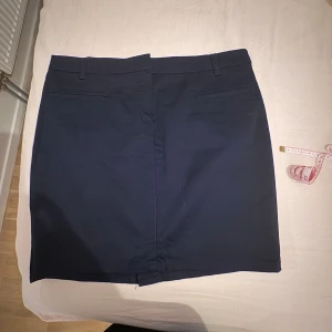 Mörkblå kortkjol från Mango Basic - Snygg mörkblå kortkjol från Mango Basic med klassisk rak passform. Kjolen har hällor för bälte, två dekorativa bakfickor och stängs framtill. Perfekt för en clean och stilren look. Materialet är slätt och känns bekvämt mot huden.