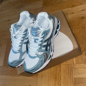 ASICS sneakers i silver och blå - Fräscha ASICS sneakers med mesh-ovandel i vitt, detaljer i silver och ljusblått samt svarta inslag. Skorna har snörning, Ortholite innersula och en chunky platt sula med blå och vita inslag. Perfekt för dig som gillar sportig och modern stil. Dem är helt nya och är slutsålda i butik.