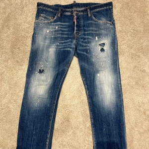DSQUARED2 blå jeans med slitningar - Snygga blå jeans från DSQUARED2 i storlek 52 med coola slitningar och färgstänk på både fram- och baksida. Klassisk femficksmodell med logotypdetaljer och en modern, smal passform. Tillverkade i bomull med jeansmaterial för en autentisk känsla.