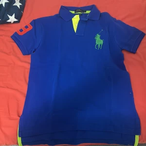 Blå pikétröja från Polo Ralph Lauren - Snygg blå pikétröja från Polo Ralph Lauren i custom fit. Tröjan har korta ärmar, grönt broderat logomärke på bröstet och gula detaljer vid knäppningen. På ena ärmen finns en röd siffra 3. Perfekt för dig som gillar klassisk och sportig stil. Jag säljer den här tröjan eftersom den är för liten och jag fick den i present därför kan jag inte skicka tillbaks den 