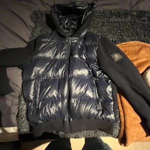 Mörkblå pufferjacka från Bvaldi - Säljer en mörkblå pufferjacka från Bvaldi med huva och snygga stickade ärmar. Jackan har två fickor med dragkedja och en cool patch på ena ärmen. Perfekt för dig som vill hålla dig varm under hösten och vintern. Nypris 2500 och använd 1 gång. Säljer för att den inte passar. Storlek m. Priset är inte hugget i sten och hör av dig vid minsta fundering eller intresse av fler bilder!