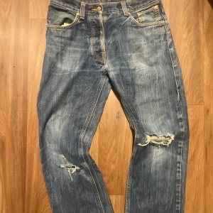 Blå Nudie Jeans W30 L32 med slitningar - Säljer ett par blåa washed jeans  från Nudie Jeans med coola slitningar Klassisk femficksmodell med orangea sömmar och snygga broderade bakfickor. Jeansen har normal passform och är tillverkade i robust bomullstyg.
