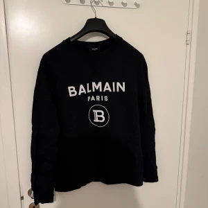 Svart sweatshirt från Balmain Paris - Svart sweatshirt från Balmain Paris med vit logotyp och tryck framtill. Tröjan har rund hals, långärmade ärmar och en klassisk passform. Materialet är mjuk bomull som känns skönt mot huden. Perfekt för dig som gillar stilrena designer och exklusiva märken. Äkta såklart 