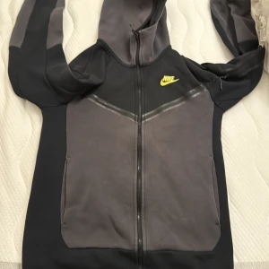Nike hoodie med dragkedja XS - Snygg hoodie från Nike i svart och grått med gul logga på bröstet. Tröjan har hel dragkedja, huva och stora fickor framtill. Perfekt för chill eller träning, med en modern och sportig look.