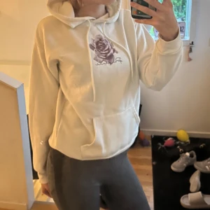 Vit hoodie med lila rostryck - Säljer en vit hoodie med lila ros-tryck på bröstet och snörning i huvan. Tröjan har en stor magficka och är långärmad, perfekt för mysiga dagar. Passformen är normal och materialet känns mjukt och skönt.