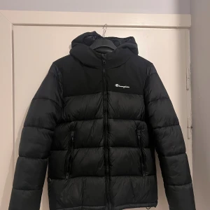 Svart pufferjacka från Champion M - Säljer en svart pufferjacka från Champion i storlek medium. Jackan har huva, dragkedja framtill och två fickor med dragkedja. Diskret Champion-logga på bröstet och på huvan. Jackan är vadderad med tech fill.