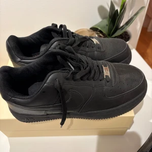 Svarta Nike Air Force 1 sneakers - Säljer ett par helsvarta Nike Air Force 1 sneakers i klassisk low-modell. Skorna har snygg skinnöverdel, perforerad tå och svarta snören med silverfärgade AF1-detaljer. Perfekta för dig som vill ha en clean och tidlös look.