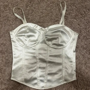Satin topp - Säljer en snygg satin korsettliknande topp. Toppen har en croppad passform, rosett framtill och band. Säljs då den inte kommer till användning. Den är väldigt sparsamt använd vilket gör att den är i nyskick.