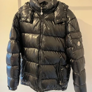 Svart pufferjacka från Moncler - Snygg svart pufferjacka från Moncler med glansig finish, hög krage och dragkedja framtill. Jackan har en ficka med lock och Moncler-logga på ärmen samt flera dragkedjeförsedda fickor. Perfekt för kalla dagar och riktigt street.
