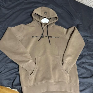 Beige hoodie från Sail Racing - Snygg beige hoodie från Sail Racing med svart logga på bröstet och huvan. Tröjan har känguruficka, svarta snören och är fodrad på insidan för extra komfort. Perfekt för dig som gillar en clean och stilren look. Änvänd få gånger