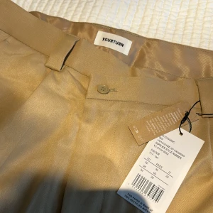 Beiga kostymbyxor - Oanvönda  - Stilrena beiga kostymbyxor från YOURTURN med rak passform och klassisk design. Byxorna har hällor för bälte, knapp- och dragkedjestängning samt diskreta bakfickor. Materialet har en lätt glansig finish som ger en dressad look.