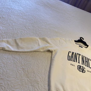 Vit sweatshirt från Gant NHCT - Vit sweatshirt från Gant NHCT med blå tryckt logga och text framtill. Klassisk rund halsringning och långa ärmar. Mjuk och skön bomullskvalitet, perfekt för en avslappnad stil. Snygg ribbad mudd vid ärmslut och nederkant.