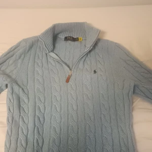 Ljusblå stickad polotröja Polo Ralph Lauren - Säljer en ljusblå stickad polotröja från Polo Ralph Lauren i klassisk kabelstickad design. Tröjan har hög krage, half zip-dragkedja och broderad logga på bröstet. Perfekt för dig som gillar stilren och tidlös look.