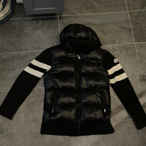 Svart pufferjacka från Moncler - Svart pufferjacka från Moncler med huva och två vita ränder på ärmarna. Jackan har dragkedja framtill och två sidofickor med dragkedja. Materialmix med glansig polyester på kroppen och stickade ärmar. Moncler-logga på vänster ärm. Och är helt ny i och perfekt  pris kan diskuteras   Funkar på m också 