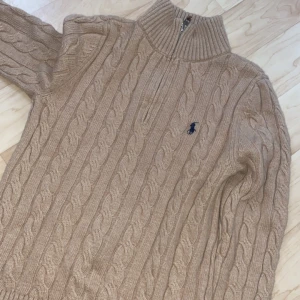 Beige kabelstickad tröja från Ralph Lauren - Kabelstickad beige tröja från Ralph Lauren med halv dragkedja och hög krage. Klassisk design med broderad logga på bröstet och långa ärmar. Perfekt för lager-på-lager och chill höst- eller vinterstil. Pris kan diskuteras vid snabb äffär