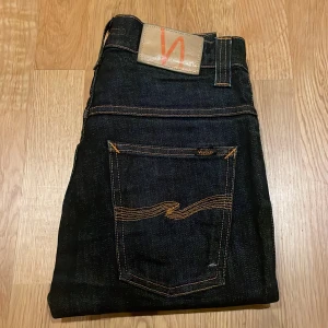 Mörkblå Thin Finn Nudie Jeans - Säljer ett par Nudie Jeans Thin Finn i mörkblå denim med klassiska orange sömmar och snygg broderad bakficka. Jeansen är tillverkade i 99% bomull och har en slim passform med normal midja. Perfekta för dig som gillar stilrena och tidlösa jeans. Pris kan diskuteras vid snabb affär!
