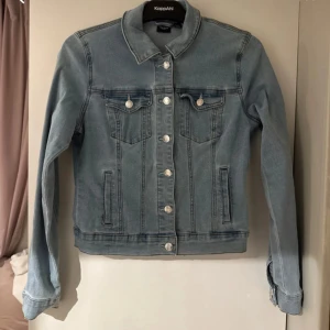 Ljusblå jeansjacka från Vero Moda - Snygg ljusblå jeansjacka från Vero Moda med klassisk krage, silverfärgade knappar och två bröstfickor med lock. Jackan har raka sömmar och lång ärm, perfekt för en chill och stilren look.