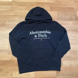 Svart hoodie Abercrombie & Fitch L - Svart hoodie från Abercrombie & Fitch i storlek L med stor vit logotyp broderad på bröstet. Tröjan har huva med snörning, magficka och långa ärmar. Storlek L!🤩