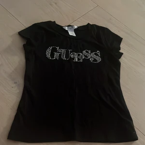 Svart t-shirt med strass från Guess barn  - Svart barn t-shirt från Guess med coolt strassmönster som bildar loggan på bröstet. Gjord i 100% bomull och har en klassisk passform med korta ärmar. Storlek XXS (4).