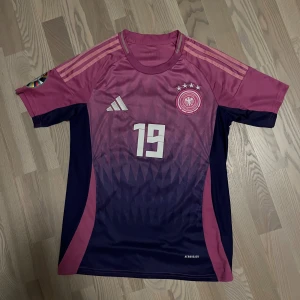 Adidas Tyskland Sané #19 fotbollströja - Säljer en officiell Tyskland fotbollströja från Adidas med Sané och nummer 19 på ryggen. Tröjan har en snygg gradient i mörkblått och rosa, korta ärmar, klassiska Adidas-ränder på axlarna och EM-märke på ärmen. Materialet är lätt och andas, perfekt för match eller träning.