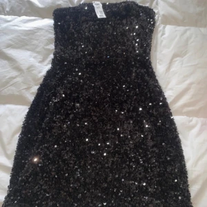 Svart paljettklänning axelbandslös - Glittrig svart axelbandslös klänning täckt av paljetter. Kort modell med figurnära passform, perfekt för att sticka ut på festen. Klänningen har en snygg, enkel siluett och är ett riktigt statement-plagg för dig som gillar att glänsa.
