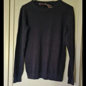 Mörkblå finstickad tröja med rund hals - En enkel och stilren mörkblå finstickad tröja med rund hals och långa ärmar. Tröjan har ribbade muddar vid ärmslut och nederkant, vilket ger en snygg passform. Perfekt för dig som gillar klassiska och tidlösa plagg. Den är lite nopprig och kan ha blivit något matt i färgen efter tvätt.