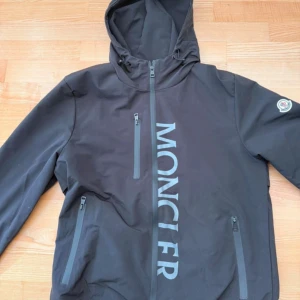 Svart vindjacka från Moncler - Svart vindjacka från Moncler med huva och dragkedja framtill. Stort Moncler-tryck längs dragkedjan och logotyp på ärmen. Jackan har två snedställda fickor med dragkedja och justerbara ärmslut. Perfekt för dig som vill ha en stilren och sportig look.