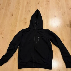 Svart hoodie från Sail Racing - Svart hoodie från Sail Racing med hel dragkedja framtill och en bröstficka med logotyp. Tröjan har huva med snörning och långa ärmar. Perfekt för chill dagar och enkel att matcha med jeans eller joggers.
