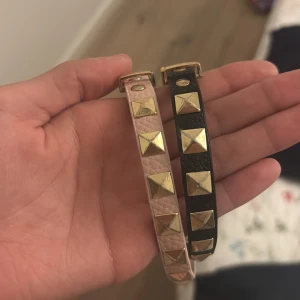Armband med nitar i guld, 2-pack - Två smala armband i läder, ett i ljusrosa och ett i svart, med coola fyrkantiga guldfärgade nitar längs hela bandet. Båda har guldfärgat spänne och detaljer. Perfekt accessoar för att ge din outfit en edgy vibe.
