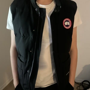 Svart dunväst från Canada Goose XS - Svart dunväst från Canada Goose i modell Fusion XS. Västen har klassisk Canada Goose-logga på bröstet, dragkedja och tryckknappar framtill samt två stora fickor. Perfekt för lager-på-lager och riktigt snygg till streetwear.