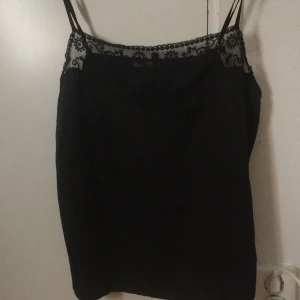 Svart linnetopp med spets från H&M - Svart linnetopp från H&M i storlek 175 med tunna axelband och en snygg spetskant upptill med blommigt mönster. Toppen är ärmlös och har en loose passform, perfekt för att ge en chill och stilren vibe. Materialet känns lätt och mjukt mot huden.