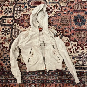 Vit quiltad hoodiejacka Parajumpers - Snygg vit hoodiejacka från Parajumpers med quiltad insida och två dragkedjeförsedda fickor fram. Jackan har ribbade muddar, huva med snörning och coolt patch på ärmen. Perfekt för dig som gillar streetwear och vill ha något som sticker ut.