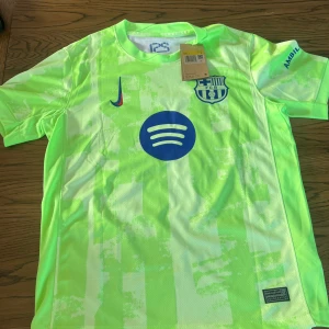 Barcelona Lamine Yamal Nike matchtröja - Barcelona matchtröja med Lamine Yamal 19-tryck, tillverkad av Nike. Tröjan är kortärmad i en ljus neongrön färg med mönster, klubbmärke och sponsorloggor i blått. Materialet är lätt och andas, perfekt för fotbollsträning eller match.