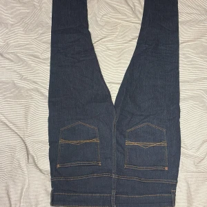 Mörkblå jeans med fickdetaljer - Snygga mörkblå jeans med raka ben och kontrastsömmar. Baksidan har två stora fickor med lock och dekorativa sömmar. Jeansen har klassisk femficksdesign och är tillverkade i ett robust denimtyg.