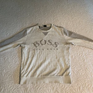 Grå sweatshirt från Hugo Boss - Snygg ljusgrå sweatshirt från Hugo Boss med stort BOSS-tryck på bröstet. Tröjan har rund hals, ribbade muddar och långa ärmar. Perfekt för en chill och stilren look. Tillverkad i mjuk bomullsmix för extra komfort.