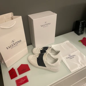 Valentino Garavani Open sneakers vit/svart - Säljer ett par snygga Valentino Garavani Open sneakers i vitt skinn med bred svart rem över foten och platt sula. Ikoniska nitar längs hälen och diskret logga på tungan. Kommer med originalkartong, dustbag och certifikat. Perfekt för dig som gillar lyxiga detaljer.